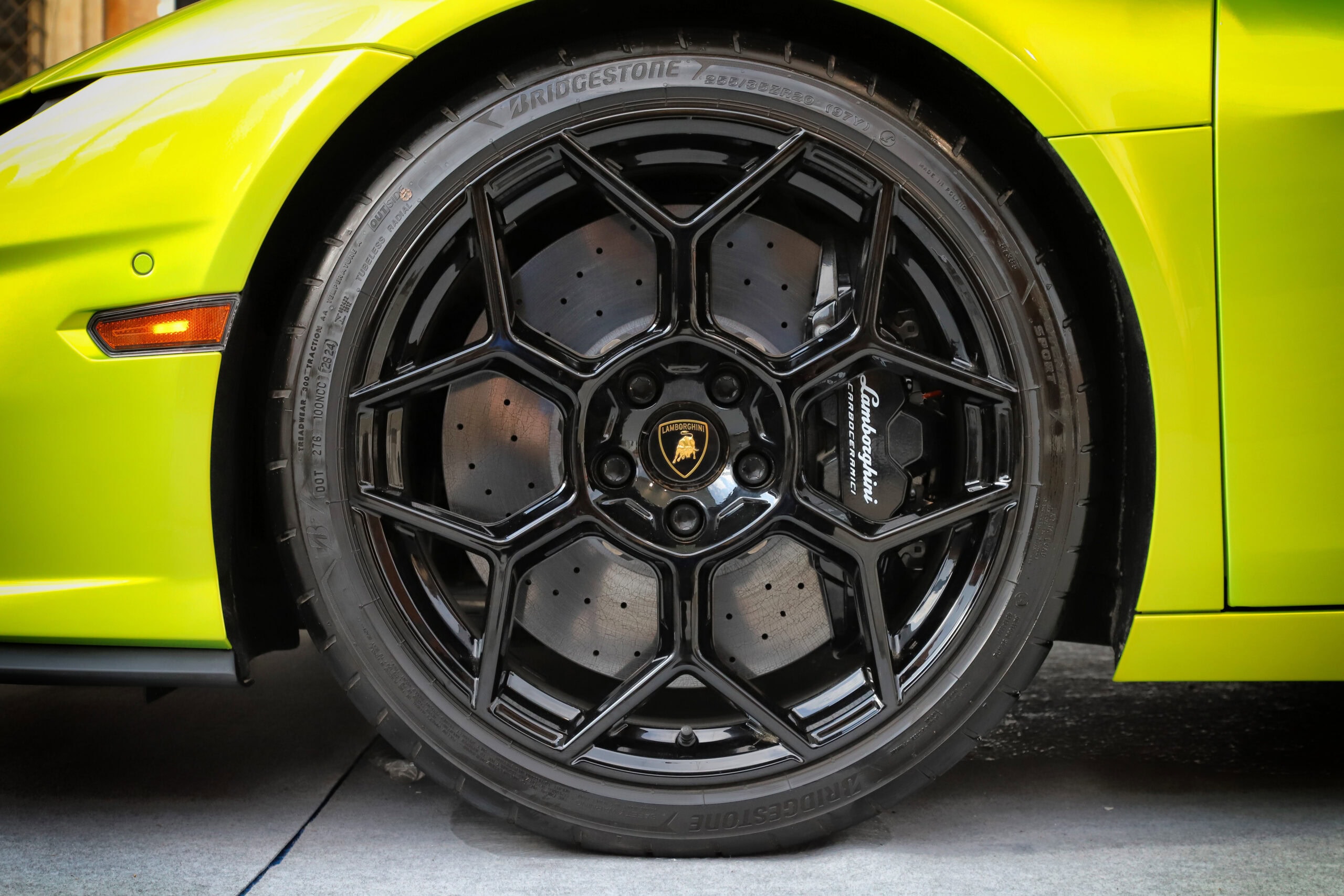 Close-up of a black lamborghini wheel with a bridgestone tire, captured at a body shop, with the yellow-green car body in the background.