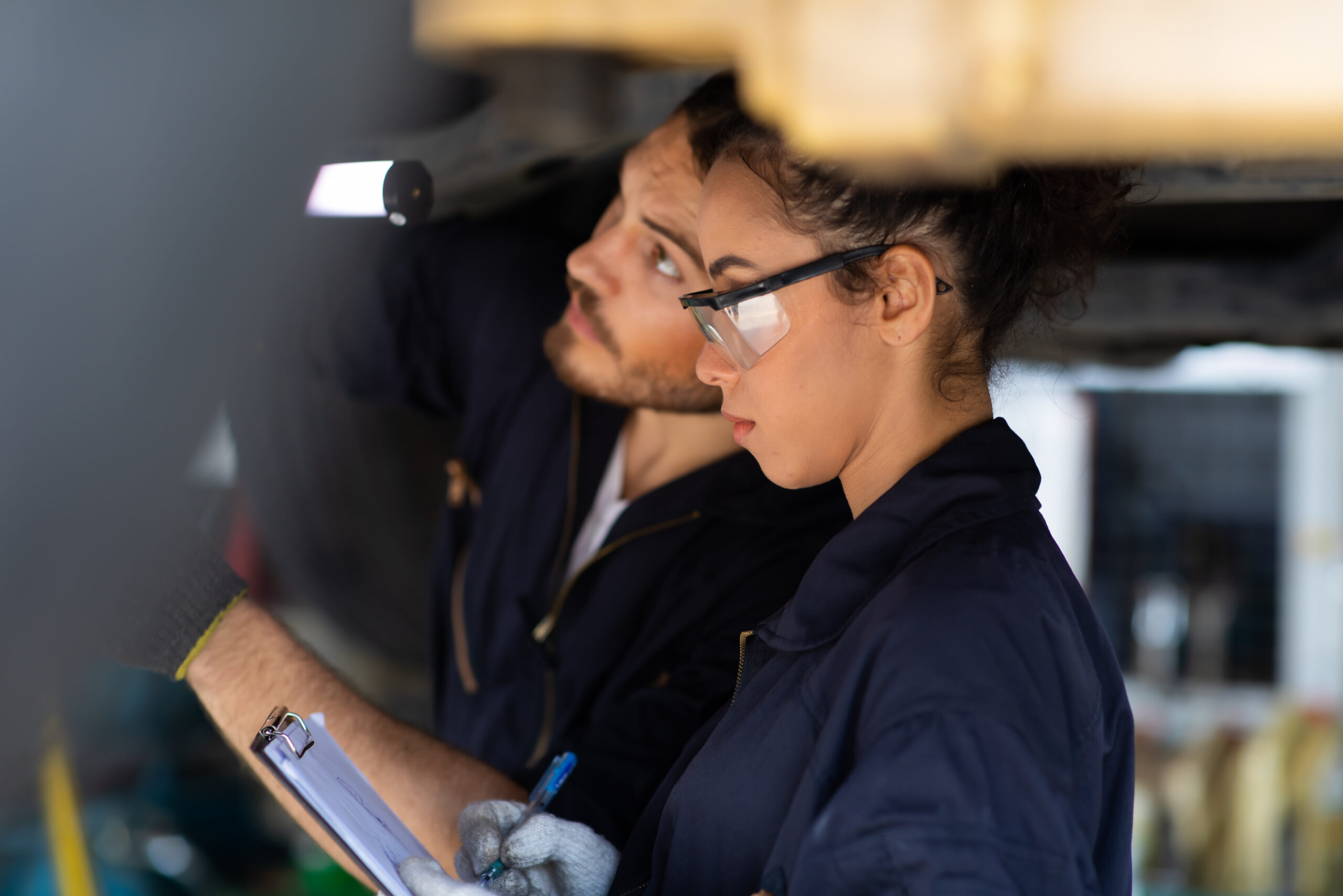 Two mechanics wearing safety glasses inspect a vehicle underside; one holds a clipboard and pen, appearing focused on taking notes.